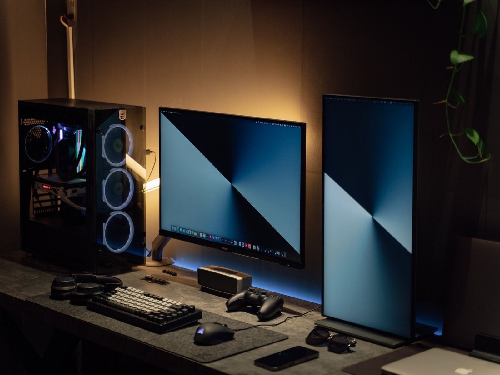 Warmly lit desk setup with a PC tower featuring blue RGB fans, dual widescreen and portrait monitors displaying blue gradient wallpapers, a mechanical keyboard, mouse, headphones, game controller, and smartphone.